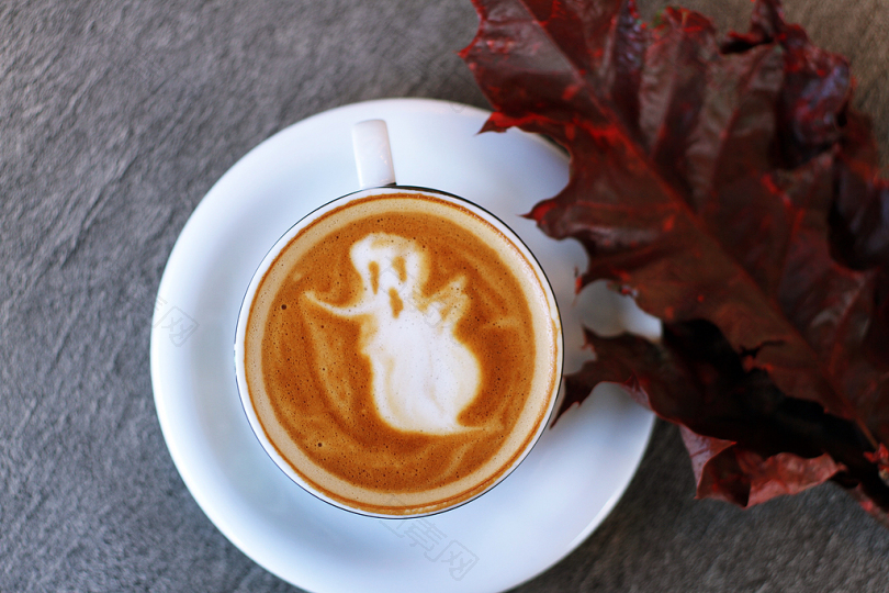 白色陶瓷茶杯充满幽灵插图咖啡拿铁在白色陶瓷茶碟旁栗色树叶摄影